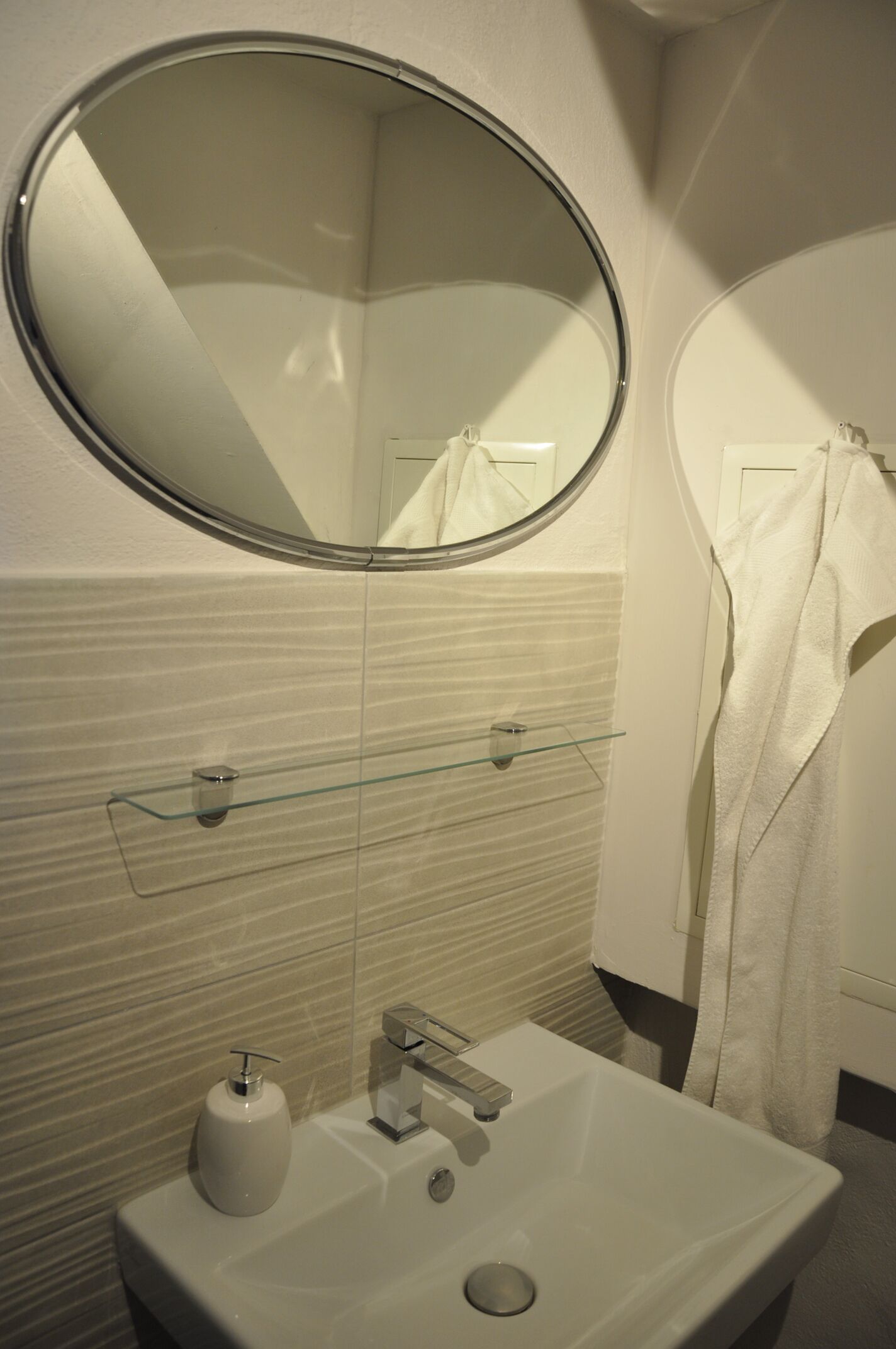 A bathroom with a round mirror, a sink, a glass shelf, and a towel rack.