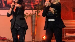 Two women on stage with microphones, a poster in the background, and a large vase with flowers. - Geibelhof (Karben)