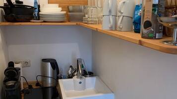 Eine Küchenzeile mit Kaffeemaschine, Wasserkocher und elektrischem Wasserkocher. Regale mit Tellern, Gläsern und einer Kaffeedose. Ein gerahmtes Foto hängt an der Wand.