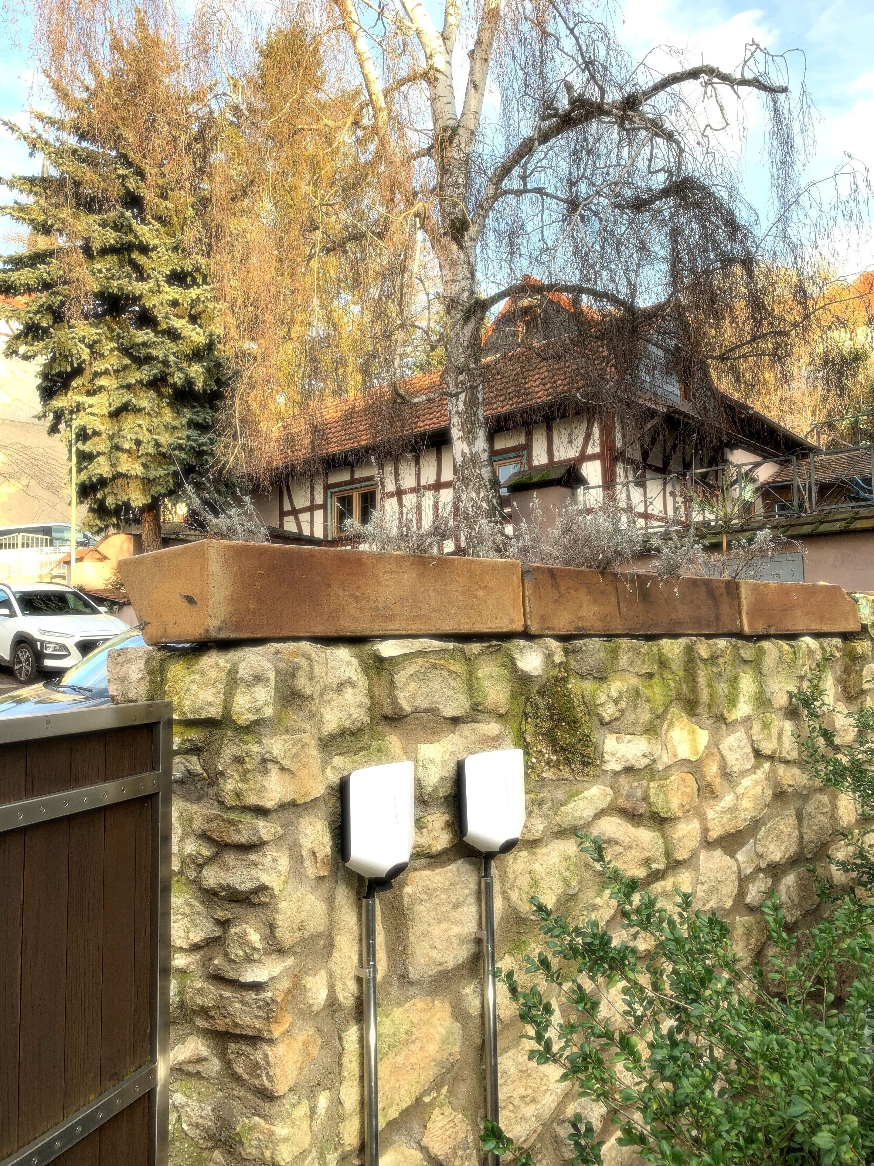A stone wall with two white objects mounted on it, a wooden gate, and a building with a tiled roof in the background.