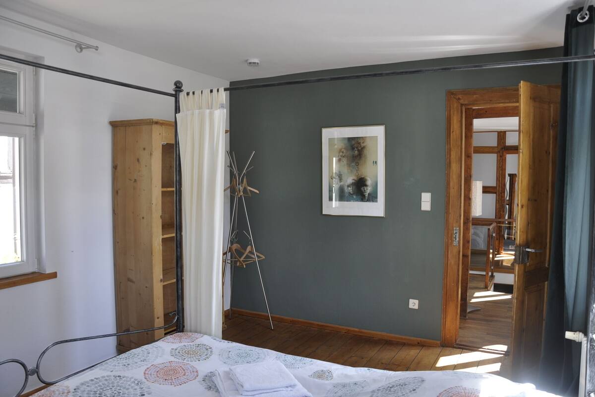 A bedroom with a bed, a wooden wardrobe, a framed picture on the wall, and a door leading to another room.