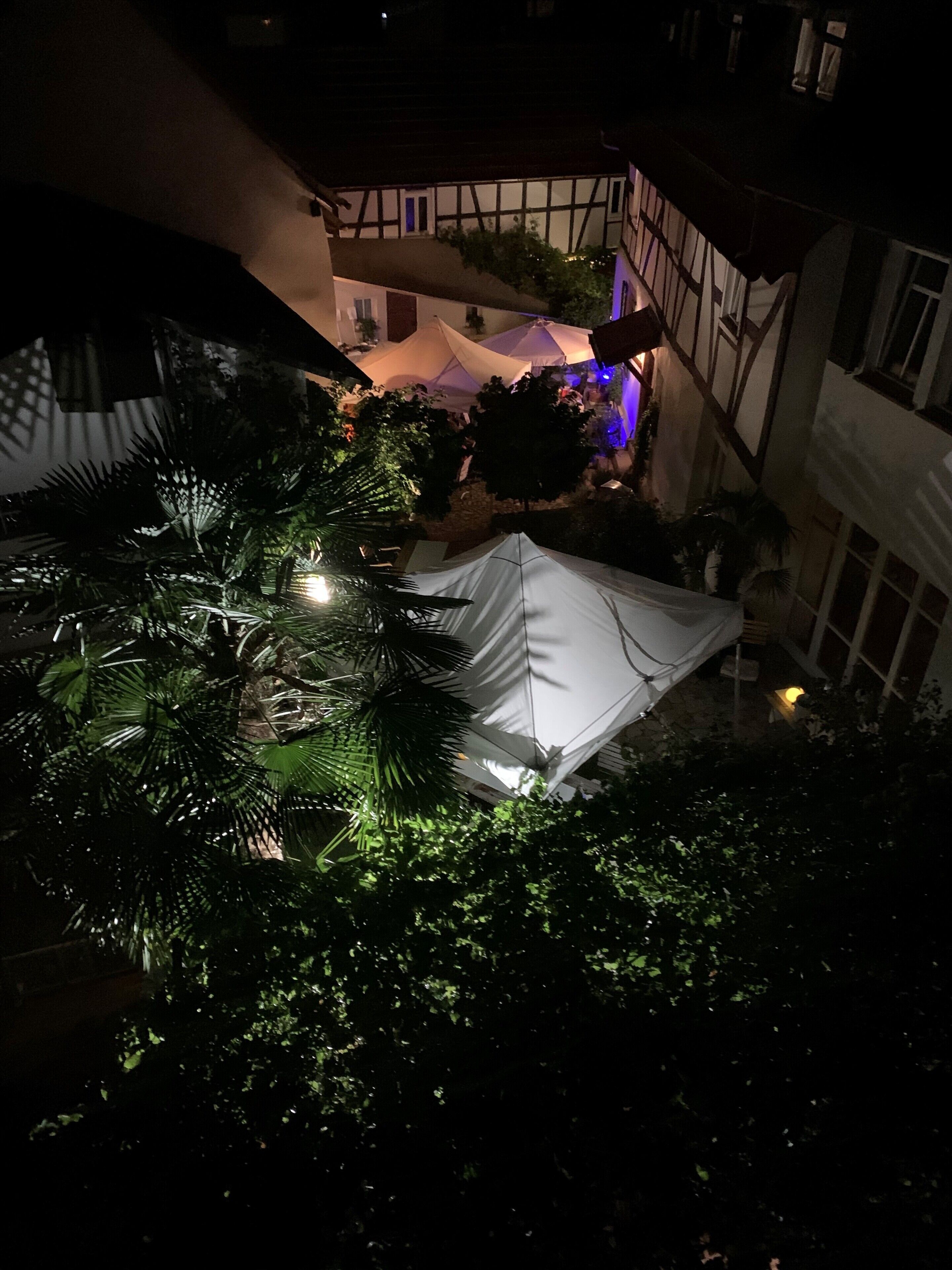 A courtyard with a tent, surrounded by lush greenery and a building with exposed wooden beams.