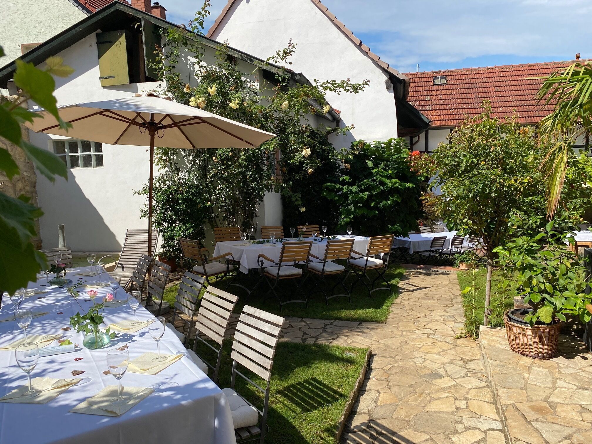 Un área de comedor al aire libre con mesas dispuestas para una comida, rodeada de vegetación y un edificio blanco con tejado de tejas rojas.