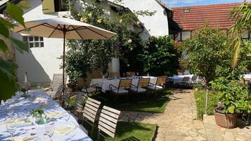 Un área de comedor al aire libre con mesas dispuestas para una comida, rodeada de vegetación y un edificio blanco con tejado de tejas rojas.