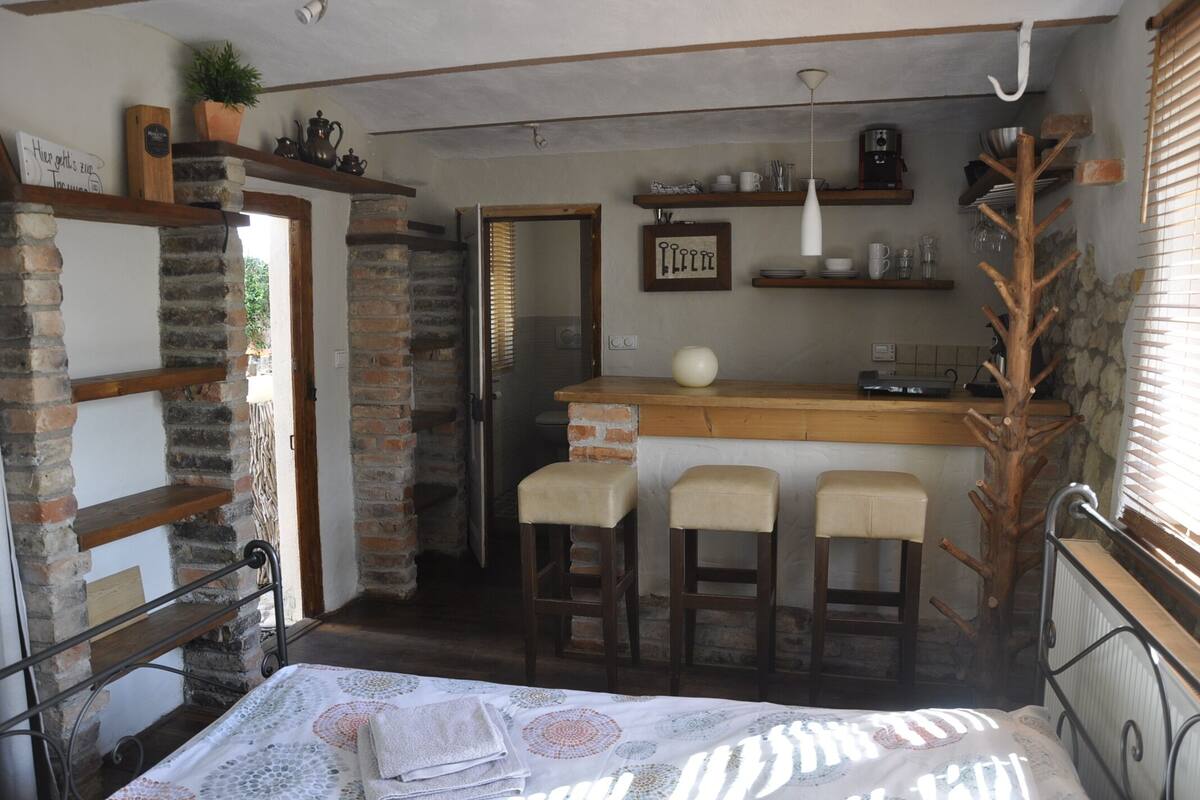 A cozy, rustic room with a bed, a wooden bench, and a small kitchen area with a brick wall and wooden shelves.
