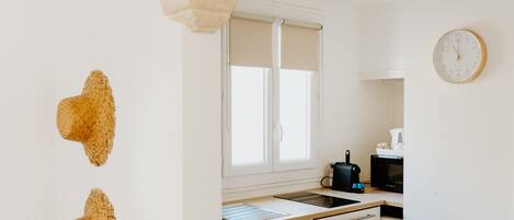 A modern kitchen with white cabinets, a black trash bin, a clock, and a window with blinds.