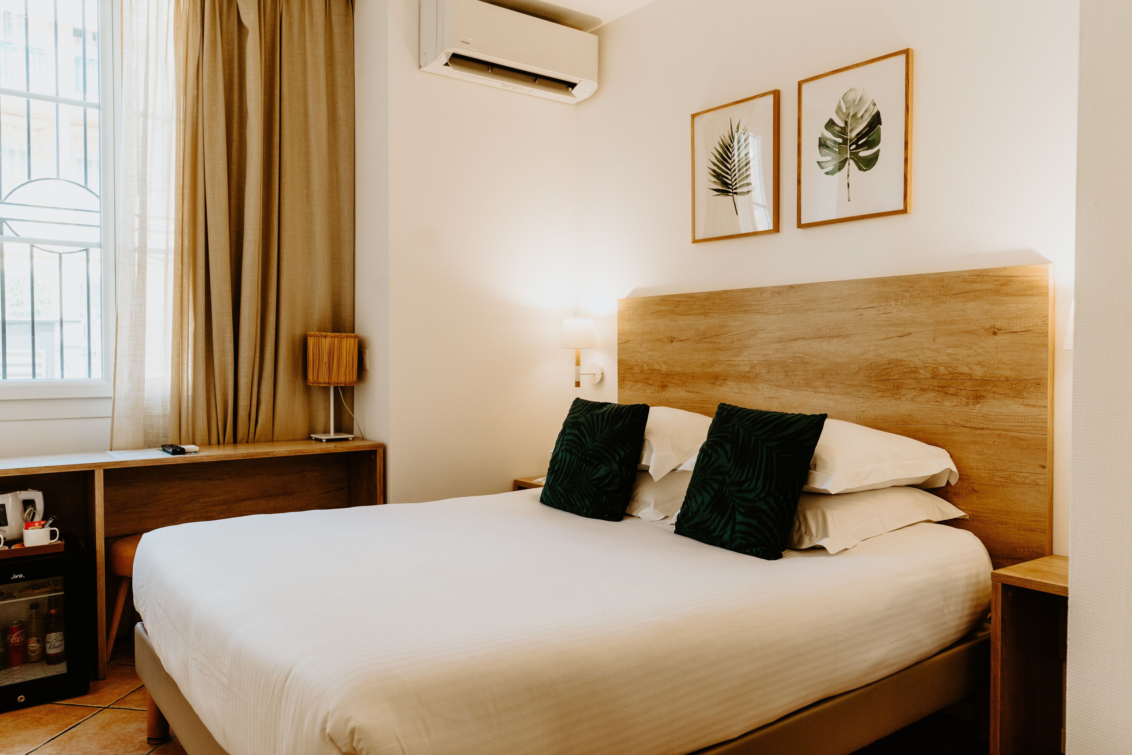 A neatly made bed with a wooden headboard, a bedside table with a lamp, and two framed botanical prints on the wall.