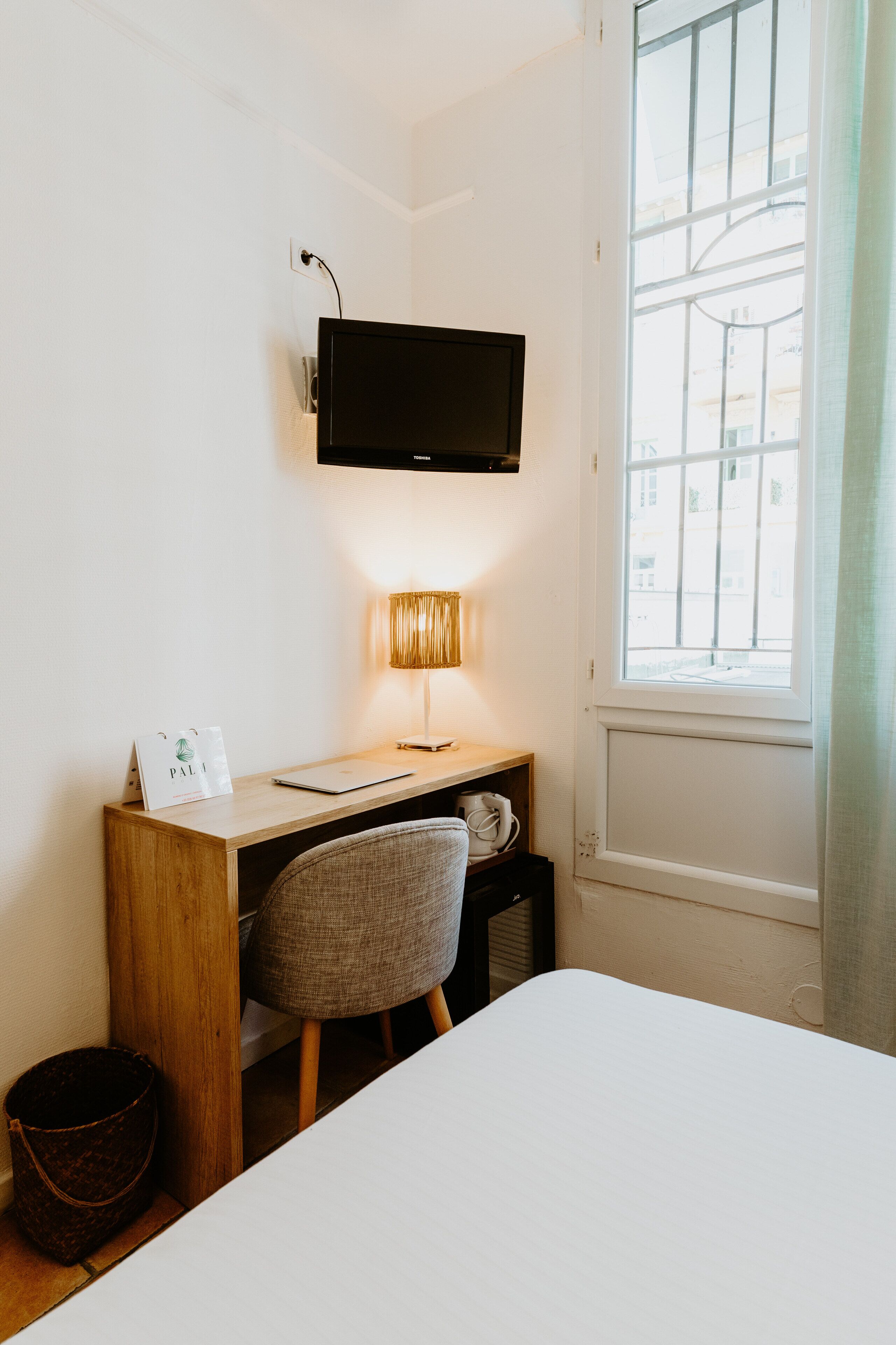 A neatly arranged hotel room with a desk, chair, and a bed. A flat-screen TV is mounted above the desk, and a lamp is placed on the desk.