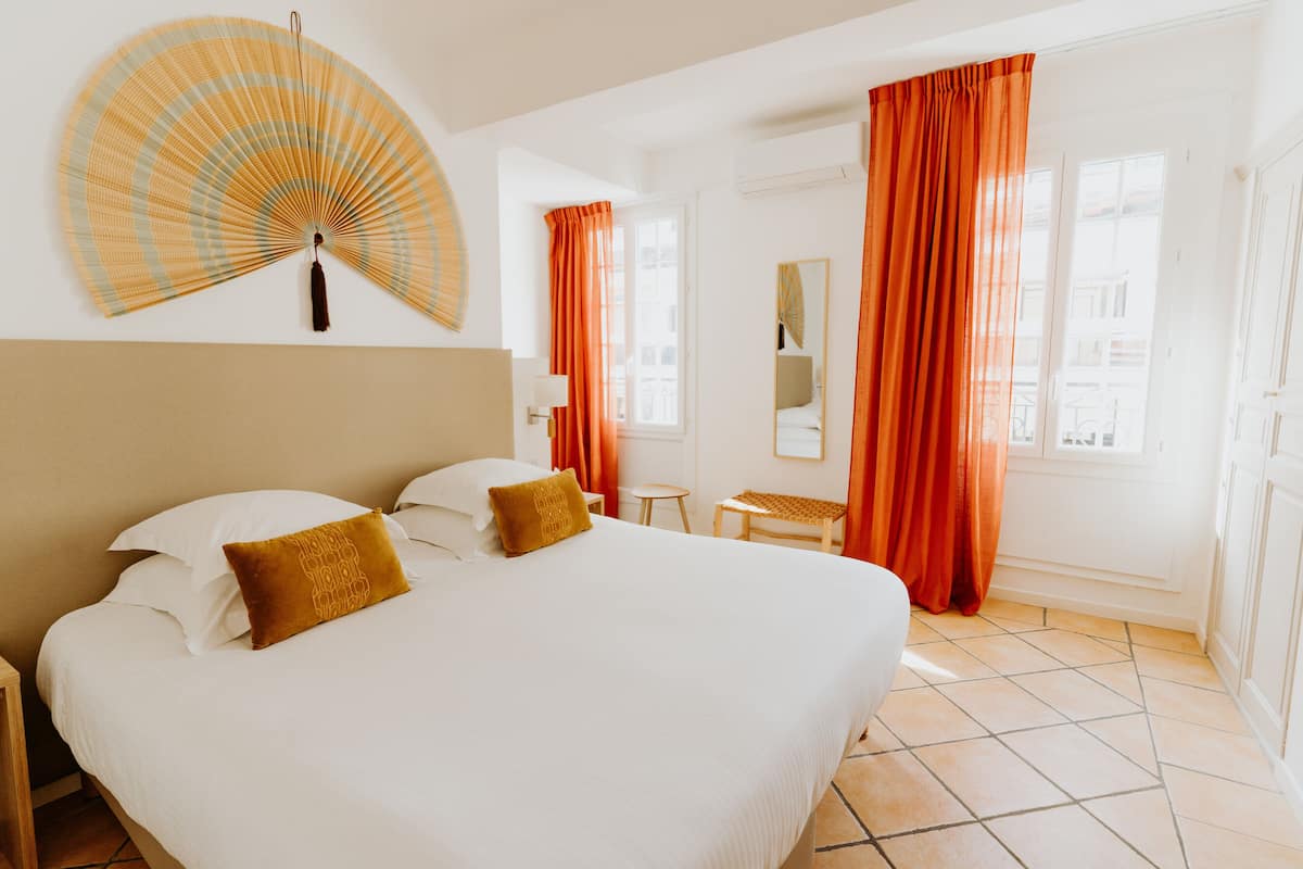 Une chambre d’hôtel avec un grand lit, deux oreillers, une table de chevet, une chaise, une petite table, un miroir et une fenêtre avec des rideaux.
