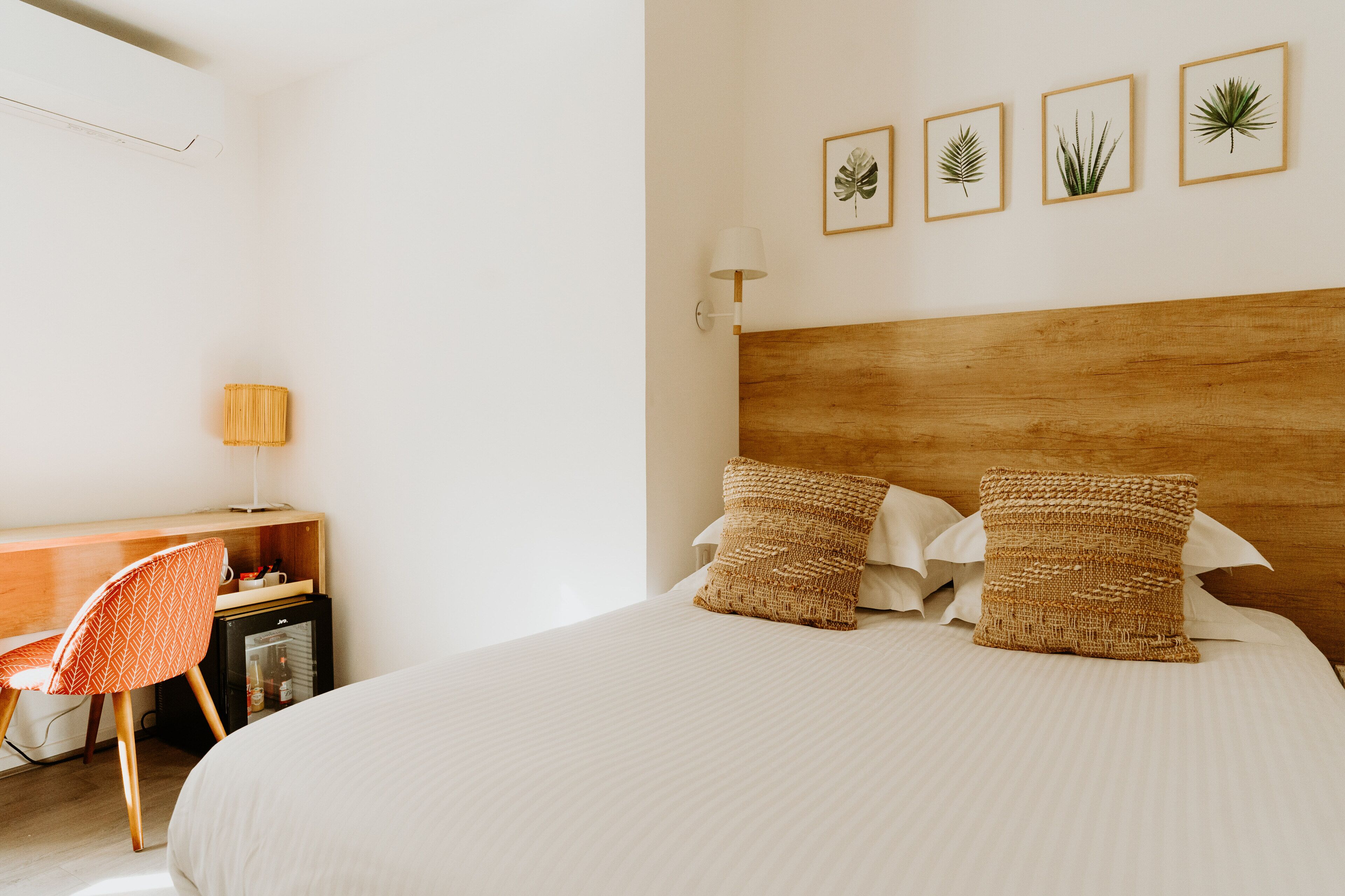 A bedroom with a wooden headboard, two woven pillows, a desk with a chair, and a wall-mounted lamp.