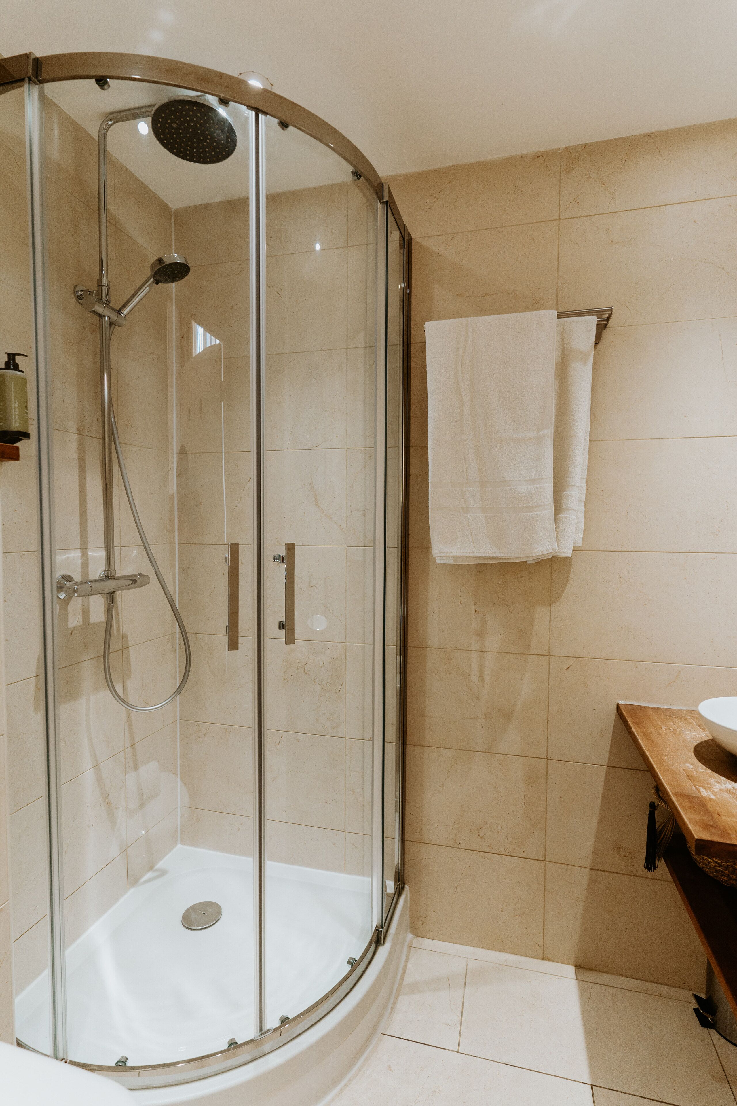 A glass-enclosed shower with a handheld showerhead, a white towel hanging on the wall, and a wooden vanity with a sink.