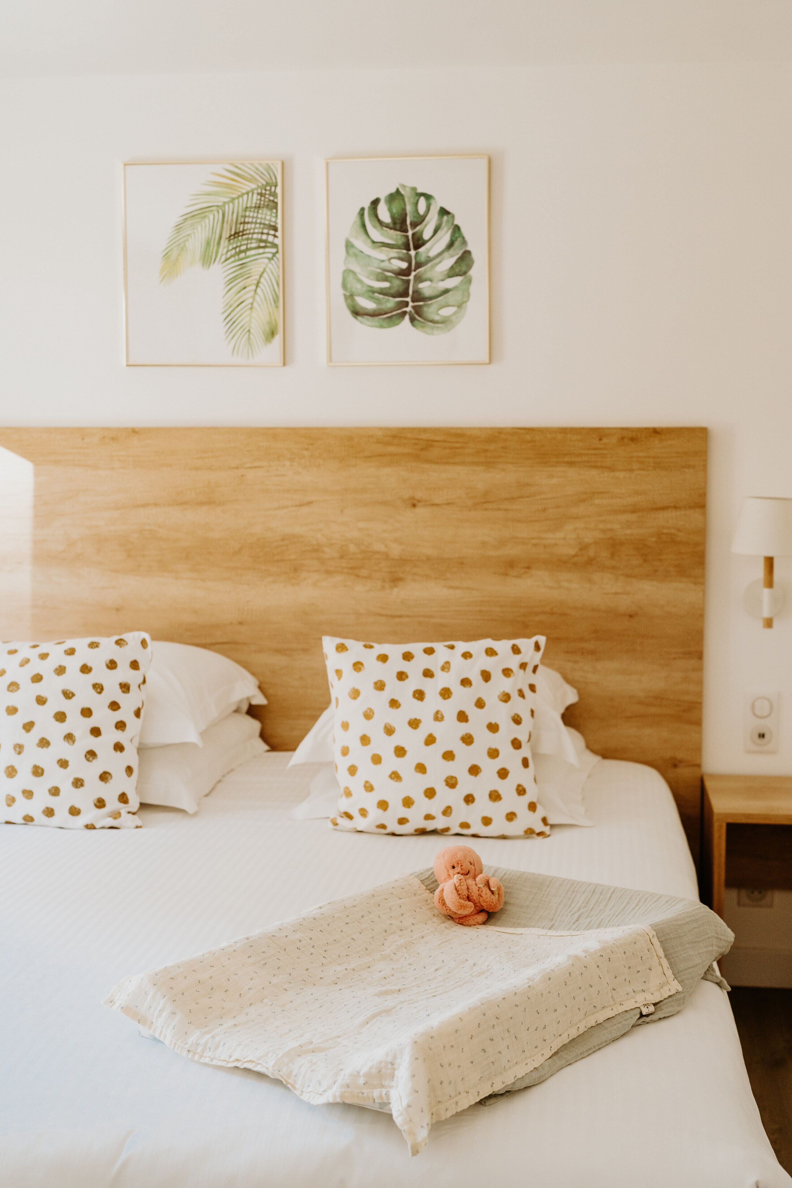 A neatly made bed with white bedding and polka dot pillows, a wooden headboard, and two framed botanical prints on the wall.