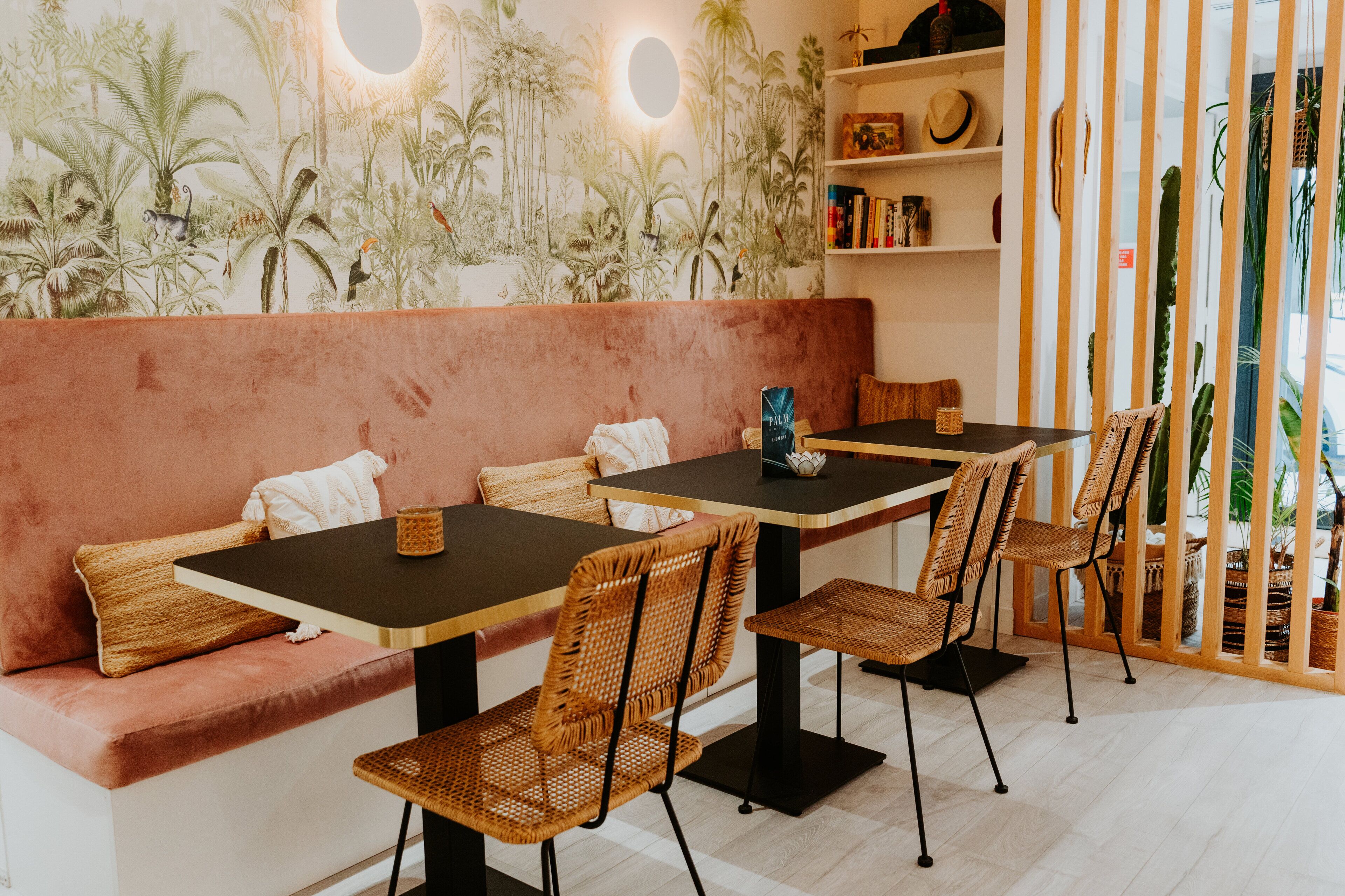 Un restaurante acogedor con un banco acolchado rosa, sillas de mimbre y un mural de plantas tropicales.
