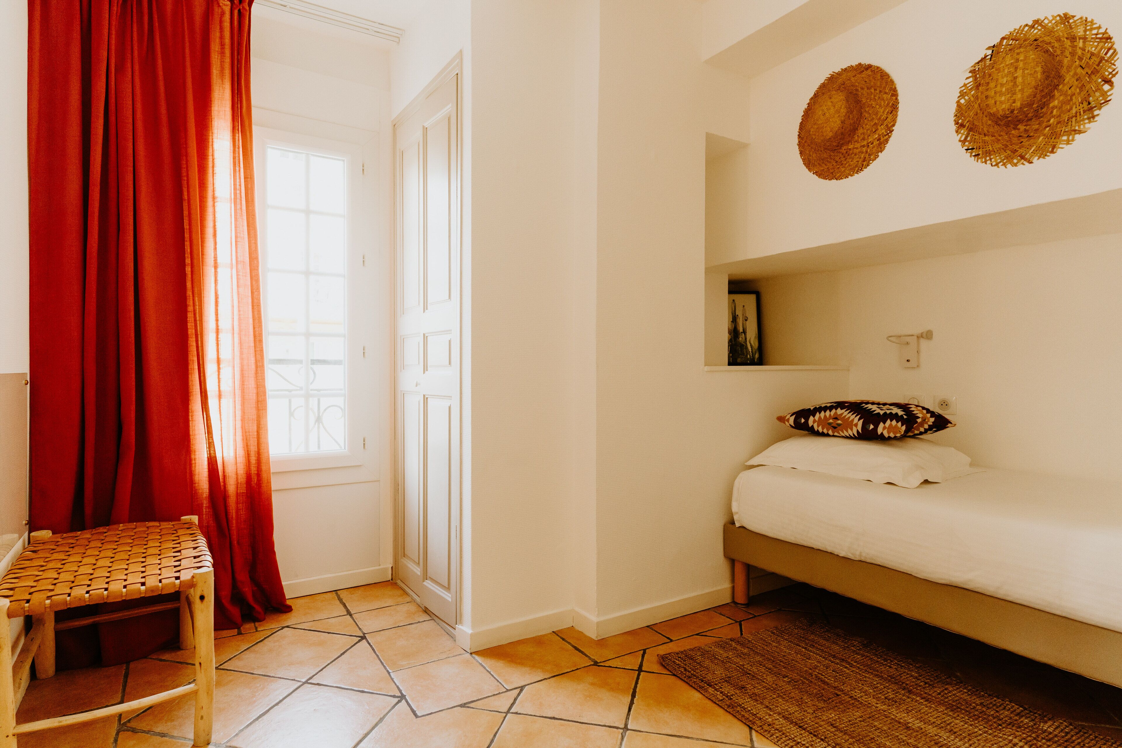 Een kamer met een bed, een stoel, een raam met gordijnen en twee hoeden aan de muur.