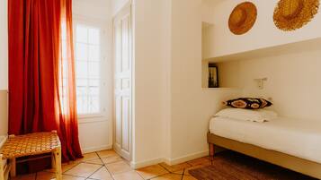 Een kamer met een bed, een stoel, een raam met gordijnen en twee hoeden aan de muur.