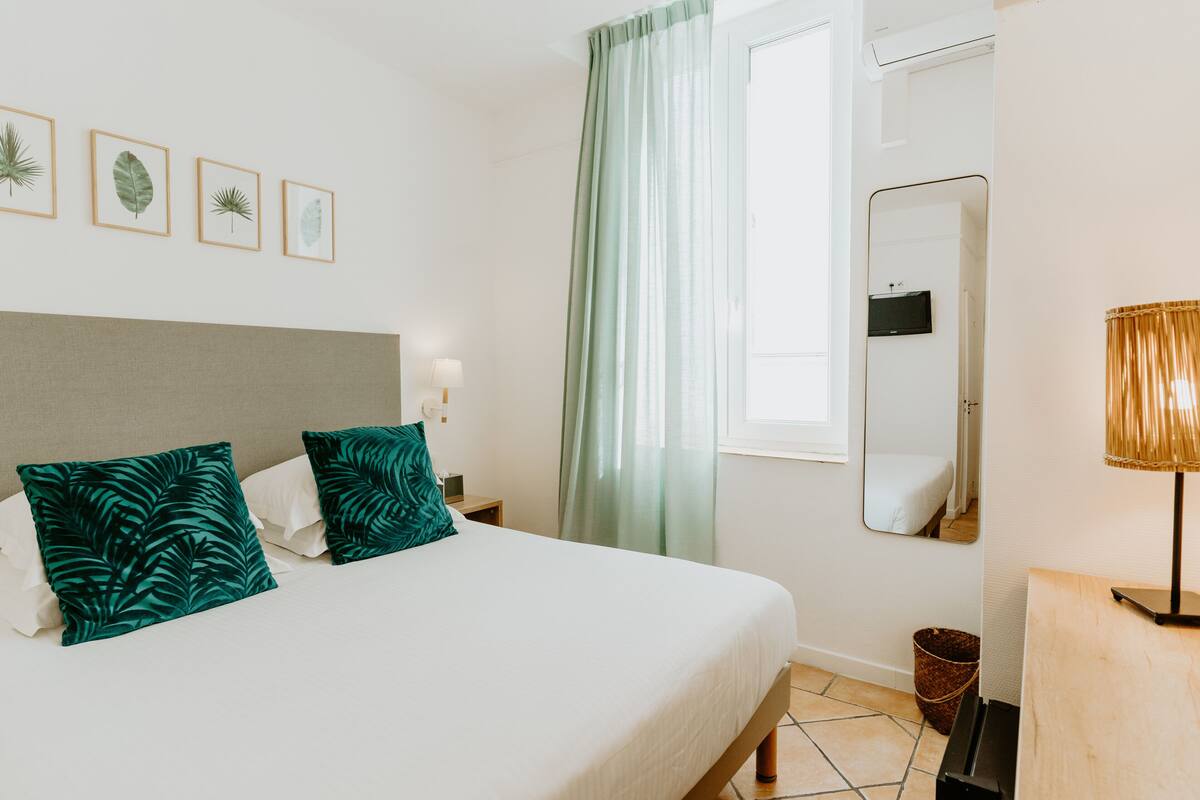 Un lit bien fait, avec deux oreillers, une table de chevet avec une lampe, un miroir et, au mur, des reproductions encadrées de motifs botaniques.