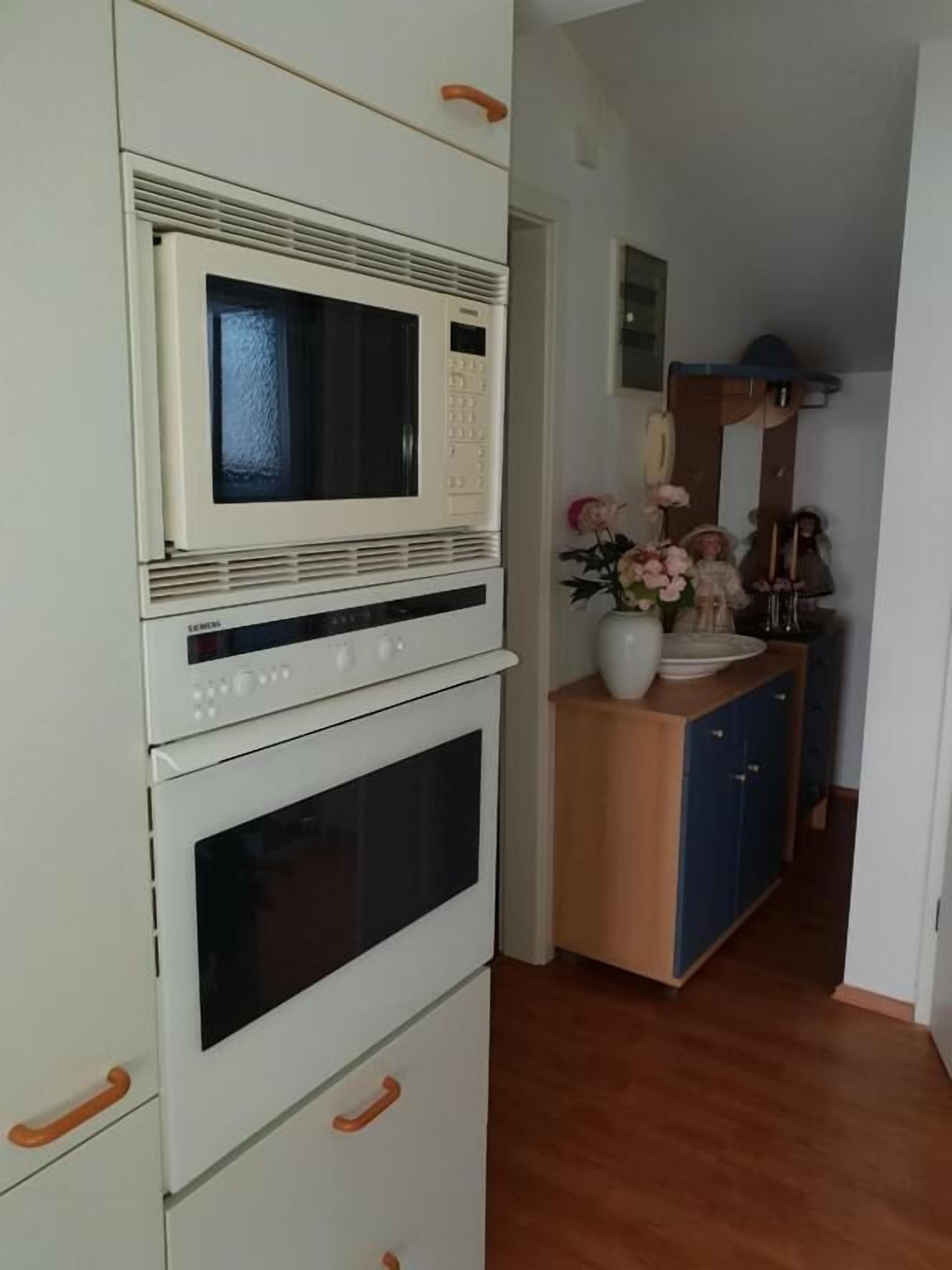 Een keuken met een magnetron, oven en kastjes. Er is een houten kastje met een bloemenvaas.