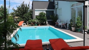 A swimming pool with red lounge chairs, surrounded by potted plants and a covered patio area.
