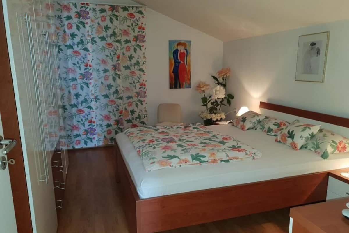 Un dormitorio con cama de madera, cortinas florales, una silla, una mesita de noche y dos cuadros colgados en la pared.