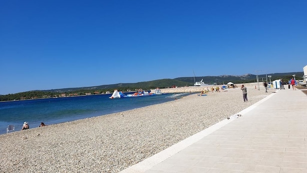 A pebbled beach with a clear blue sky, a paved walkway, and boats in the water.