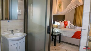 A hotel room with a glass shower enclosure, a bedside table with a lamp, a bed with white bedding and red accents, and a framed picture on the wall. - Mini Apartment at Peo (Maun)