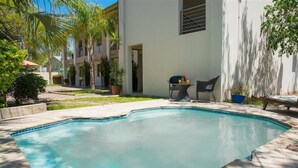 A swimming pool with a tiled deck, surrounded by potted plants and a building with a door and window. - Mini Apartment at Peo (Maun)