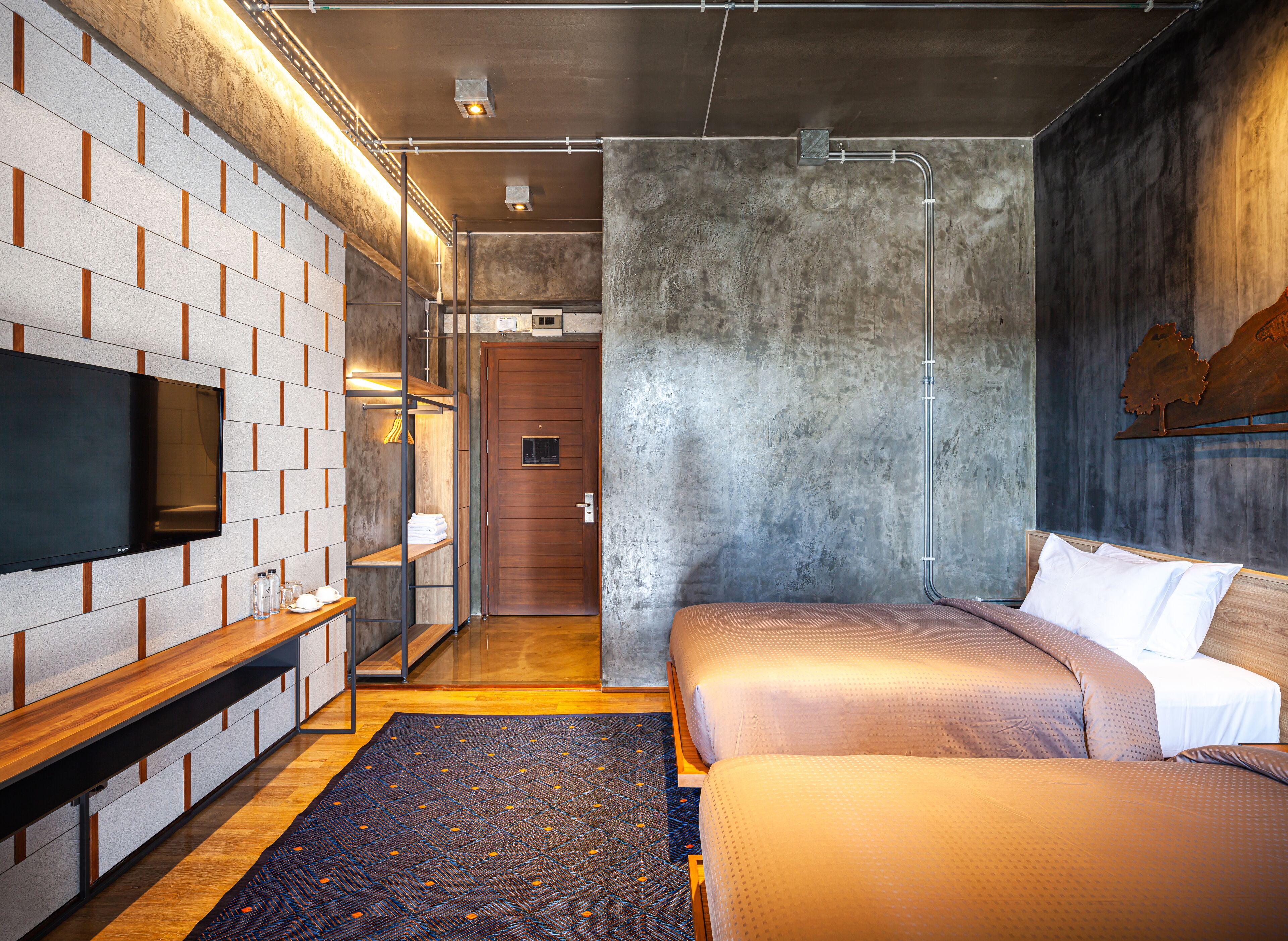 A modern hotel room with a brick-patterned wall, a flat-screen TV, a wooden bench, a large bed, and a rustic decor element.