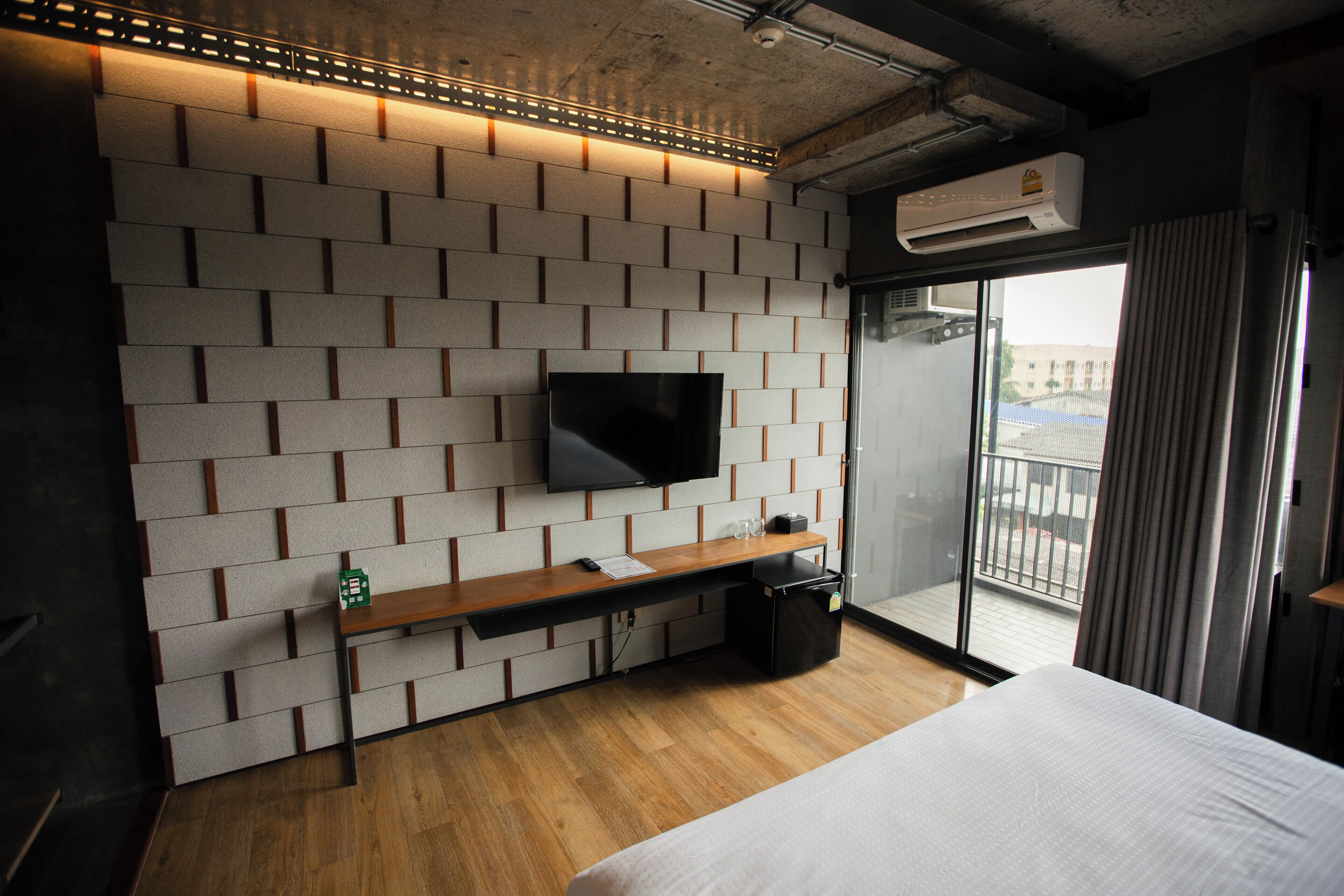 Une chambre d’hôtel moderne avec un mur décoré d’un motif en briques, un téléviseur à écran plat, un bureau en bois, une poubelle noire et un grand lit.
