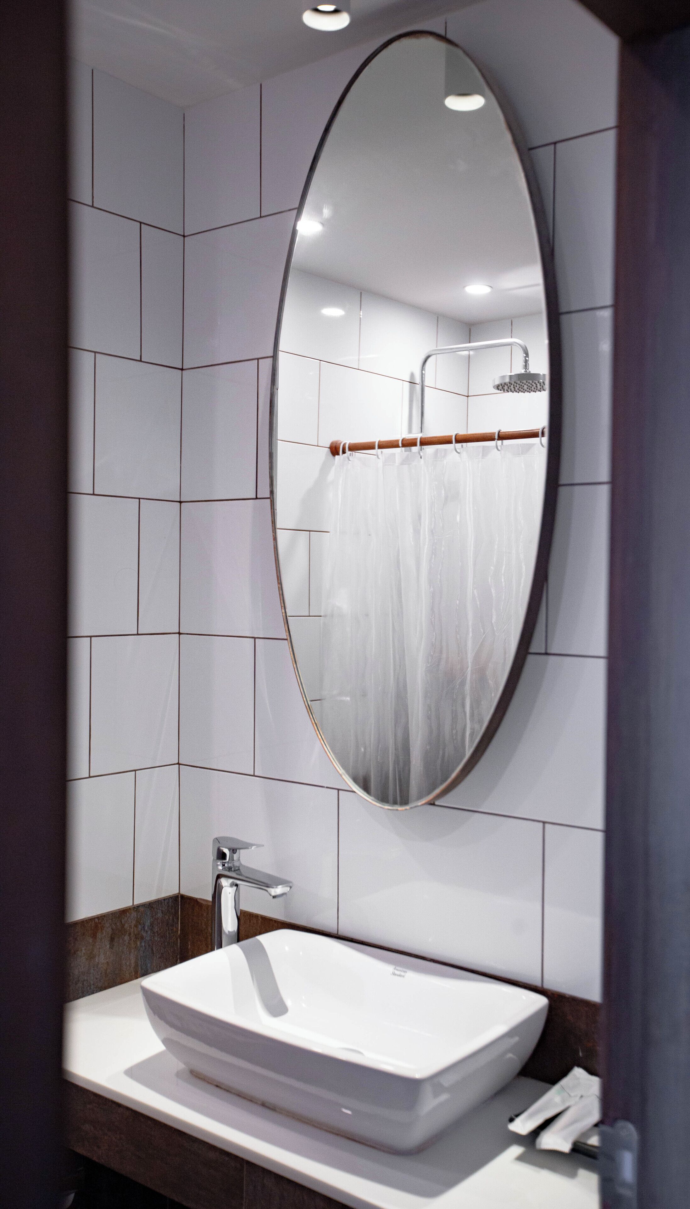 Een moderne badkamer met een grote, ovale spiegel, een wit wastafeltje en een zichtbare douchecel in de weerspiegeling.
