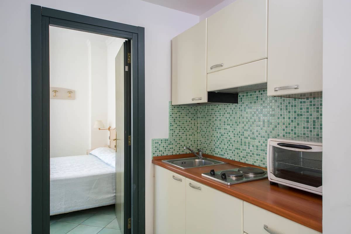 A compact kitchen with a microwave, sink, and a small dining area with a bed visible through an open door.