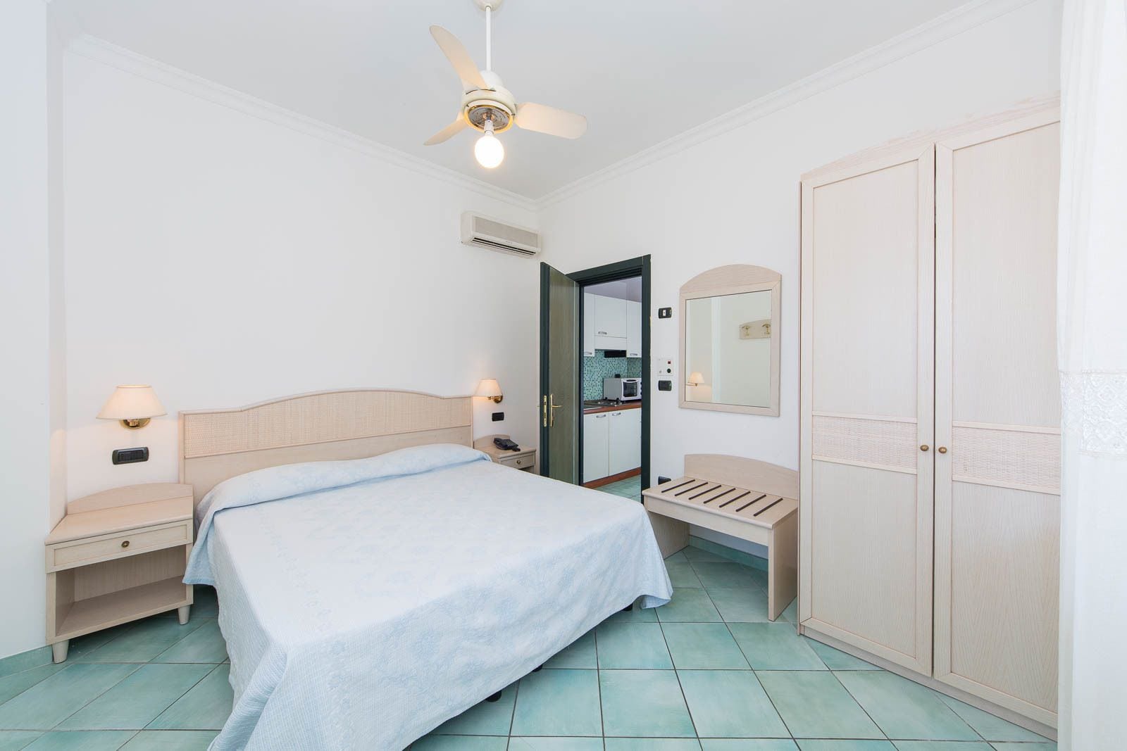 A bedroom with a bed, bedside tables, a ceiling fan, a mirror, a wardrobe, and a kitchenette in the background.