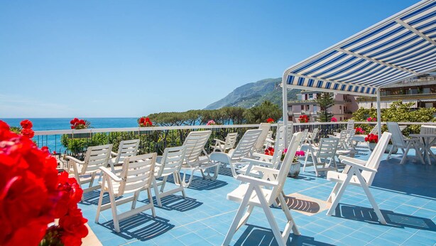 A poolside area with white lounge chairs and a covered seating area, overlooking a clear blue sea and a mountainous backdrop. - La Residenza Aparthotel (Maiori)