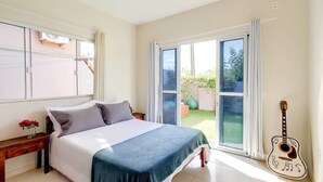 A bedroom with a bed, a nightstand, a guitar, and a view of the outside. - Origin Casa -2 Bedroom Beach House for up to 7 (Palhoça)