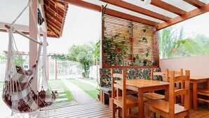 A wooden deck with a hammock, a wooden table with chairs, and a view of a garden through a window with blinds. - Origin Casa -2 Bedroom Beach House for up to 7 (Palhoça)