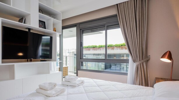 A hotel room with a large bed, a TV mounted on the wall, a window with curtains, and a bedside lamp. - Na85b Modern Studio in the Best V. Madalena (São Paulo)