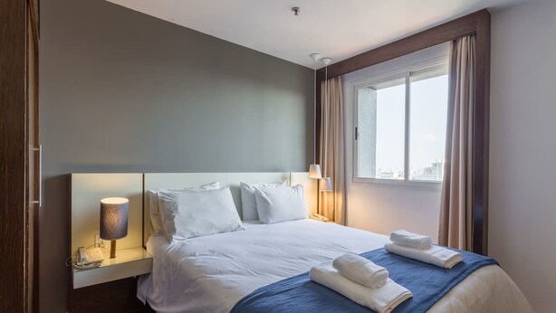A hotel room with a large bed, a bedside lamp, a window with curtains, and a city view. - No1712 Fantástico a Poucos Metros da Paulista! (São Paulo)