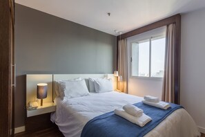 A hotel room with a large bed, a bedside lamp, a window with curtains, and a city view. - No1712 Fantástico a Poucos Metros da Paulista! (São Paulo)