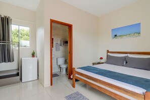 A bedroom with a bed, a small bedside table, a nightstand, a window with curtains, a radiator, and a door leading to a bathroom. - S5 Origin - Nice Suite With Balcony, View and AC (Palhoça)