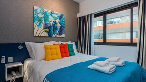 A hotel room with a bed, colorful pillows, a blue blanket, a painting, a phone, and a window with curtains. - Ad404 Stylish Apartment in the Best Part of Itaim (São Paulo)