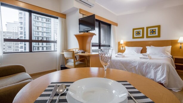 A hotel room with a bed, bedside tables, a desk, a chair, a dining table with a bowl and cutlery, and a large window with a city view. - Tu705 Varanda e Muito Conforto na Al Lorena! (São Paulo)