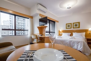 Hotellihuone, jossa on sänky, yöpöydät, työpöytä, tuoli, ruokapöytä kulhoineen ja ruokailuvälineineen sekä suuri ikkuna, josta avautuu näkymä kaupunkiin.