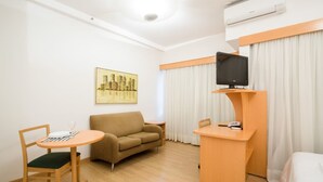 A hotel room with a sofa, a small round table with a chair, a TV on a stand, and a painting on the wall. - Tu705 Varanda e Muito Conforto na Al Lorena! (São Paulo)