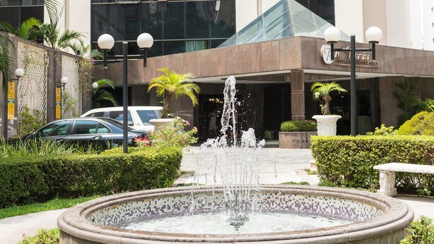 Une fontaine se trouve au centre d’un bassin circulaire bordé de carreaux, entourée de haies bien entretenues et de quelques plantes en pot. Au fond, on aperçoit un bâtiment moderne avec une façade en verre et une enseigne au-dessus de l’entrée.