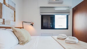 A hotel room with a bed, a desk, a lamp, a window, and a wall-mounted air conditioner. - Ad604 Estúdio a 1 Quadra da Av. Paulista (São Paulo)