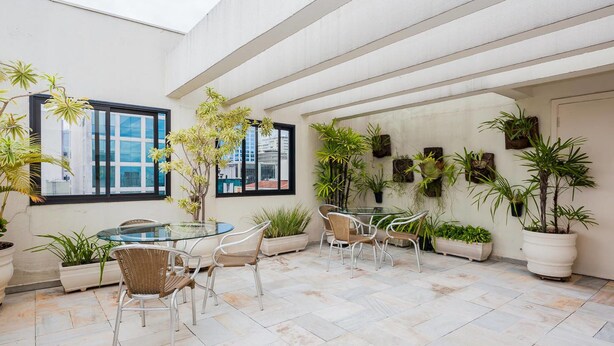 A spacious indoor patio with a glass table and chairs, surrounded by potted plants and a view of the city skyline through large windows. - Ad604 Estúdio a 1 Quadra da Av. Paulista (São Paulo)