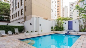 A swimming pool with lounge chairs and potted plants, surrounded by a white wall and tall buildings.