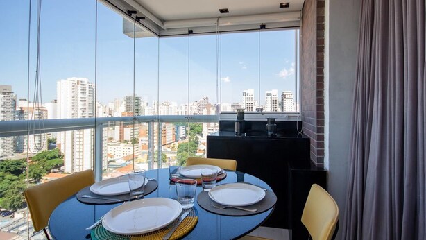 Un comedor con una mesa dispuesta para cuatro personas, ventanas amplias con vistas a la ciudad y un panel negro con una pequeña mesa y sillas.