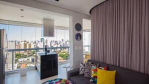 Una sala de estar moderna con un sofá, cojines de colores, una mesita redonda y vistas a un paisaje urbano a través de amplios ventanales.