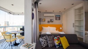 Una habitación de hotel moderna con cama, sofá, una mesita con una revista, y un balcón con mesa y sillas.