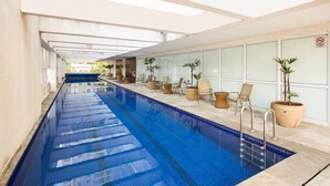 Una piscina interior larga con agua azul cristalina, rodeada de sillones de descanso y plantas en macetas.